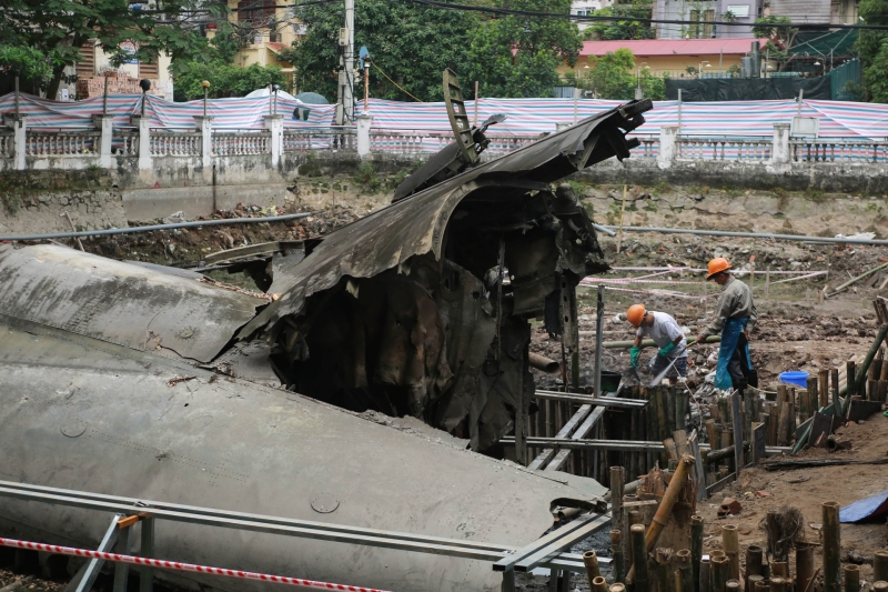 Giữa hồ nước được hút cạn, nhóm công nhân đang nạo vét bùn và quây dàn giáo quanh mảnh xác máy bay B52 để bảo dưỡng